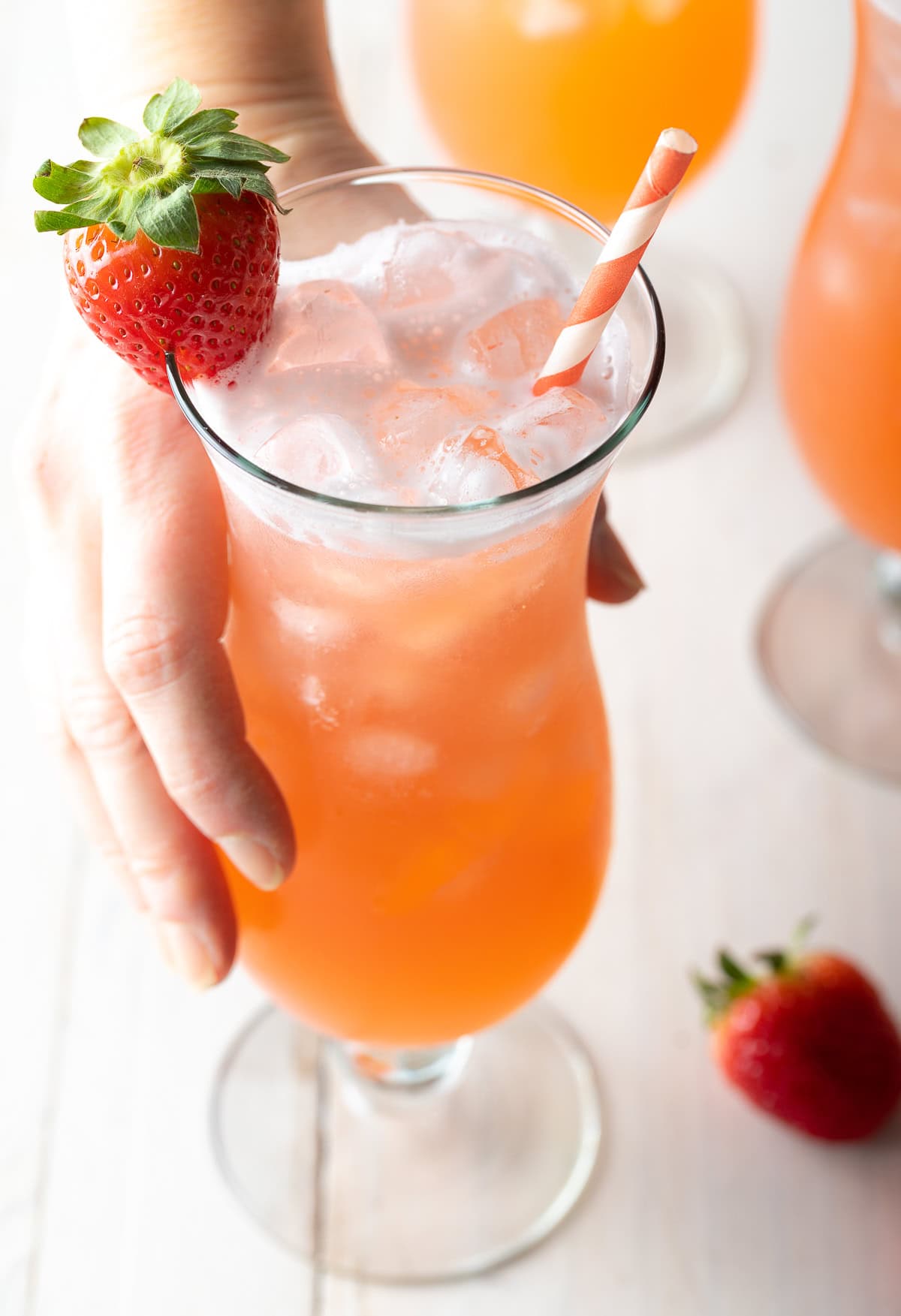 Hand holding a tall cocktail glass filled with strawberry pineapple agua fresca. The glass is garnished with a strawberry on the rim and and orange and white striped paper straw.
