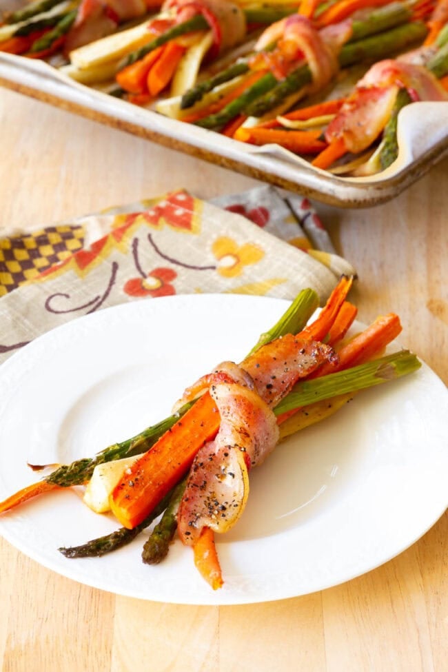 Roasted bacon wrapped vegetables on a white plate with a piece of bacon wrapped around it. 