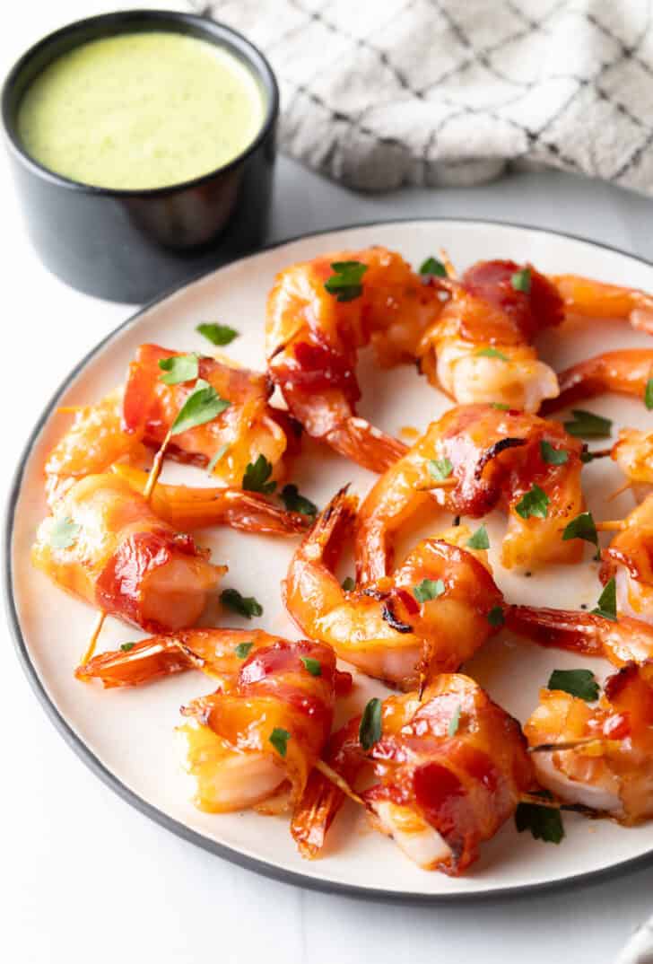 White plate with nearly a dozen bacon wrapped shrimp are loaded onto the plate in a single layer.