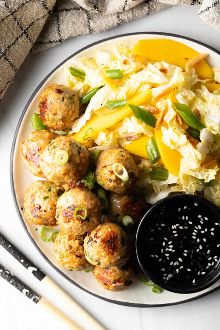 Top down view of a plate covered half Thai turkey meatballs and half chopped Asian salad, plus a black ramekin of dipping sauce.