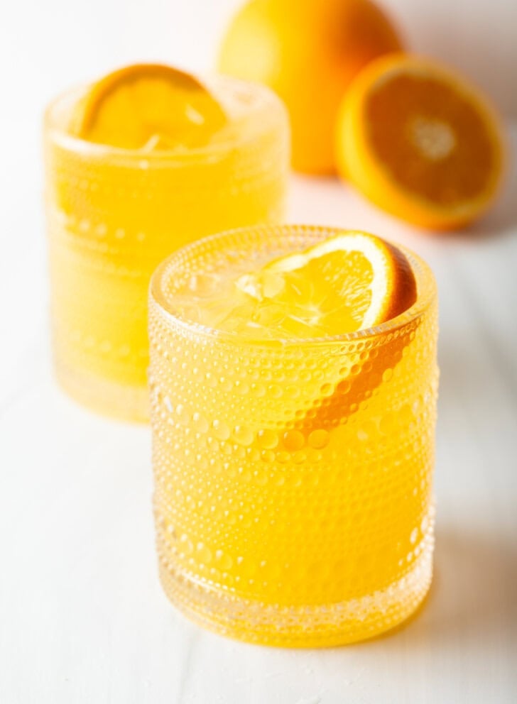 Two orange crush cocktails in glasses, each garnished with a slice of orange. There are fresh oranges and lemons around the glasses.
