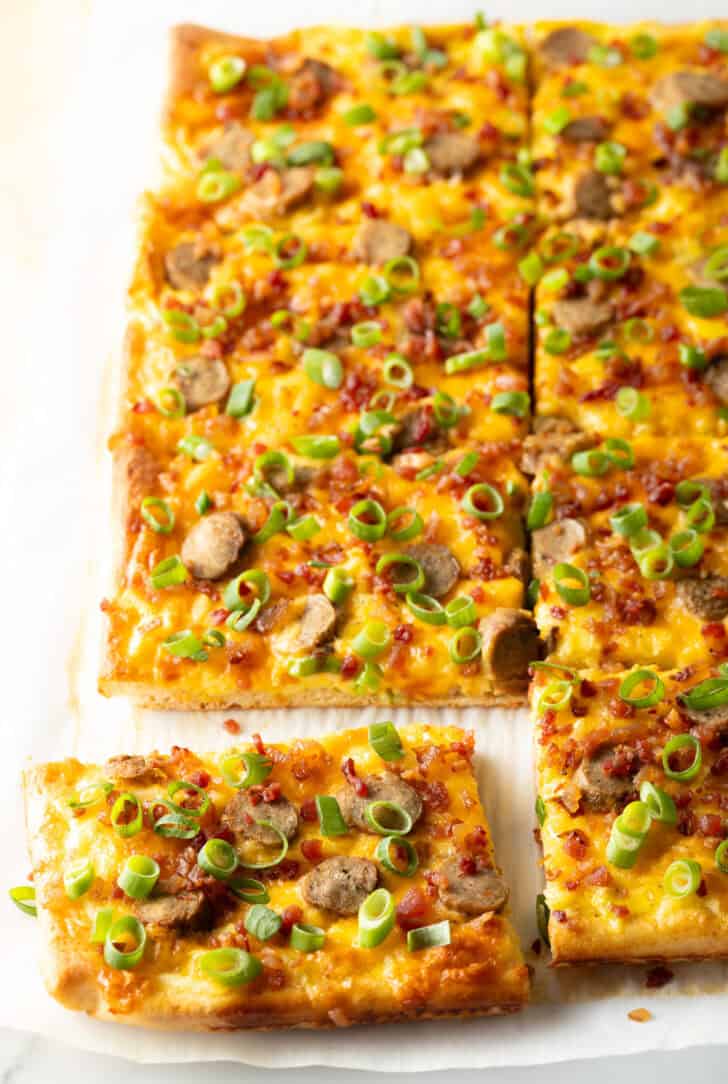 Rectangle baking sheet tray with breakfast pizza, loaded with cheese, sausage, and green onions. The pizza has been cut into 8 rectangles, with one piece pulled away.