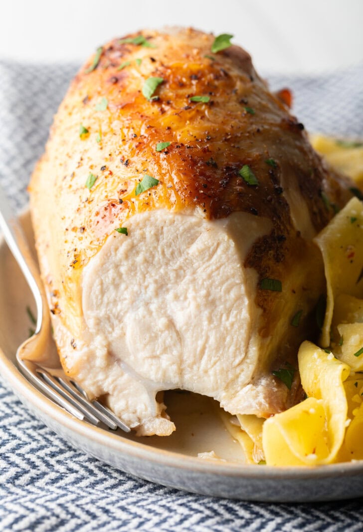 Bourbon brined chicken breast on a white plate with side of buttery noodles. A large bite has been removed from the chicken to show the moist interior.