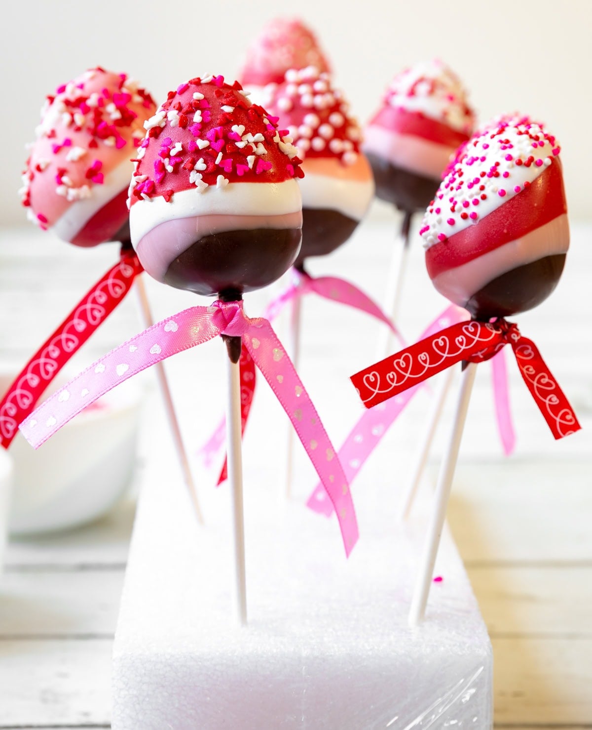 6 chocolate covered strawberry lollipops with layers of brown chocolate, pink, white, and red all topped with pink and white Valentines' day sprinkles.