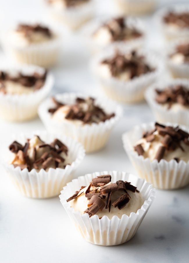 Close view several no bake keto cheesecake bites in small paper cups on a marble background with chocolate shavings on top.