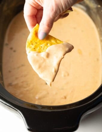 A hand holding a tortilla chip that's been dipped in crock pot queso dip, hovered over the crock pot.