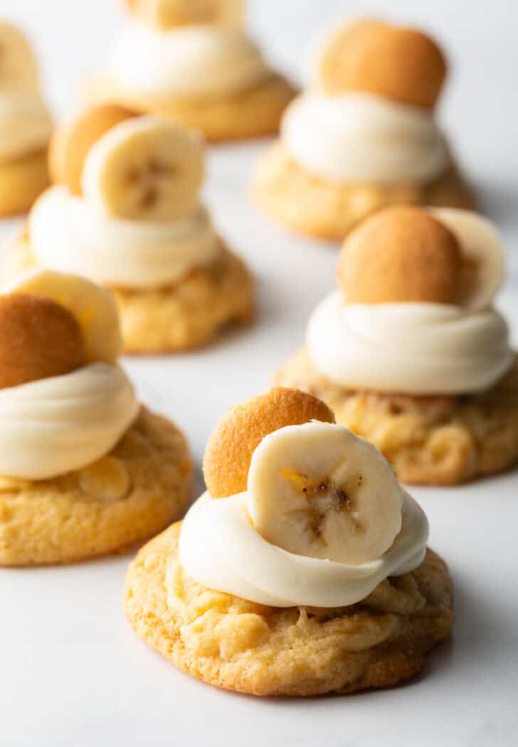 Several banana pudding cookies on a baking sheet, each topped with a dollop of vanilla frosting, a slice of fresh banana, and a Nilla wafer.