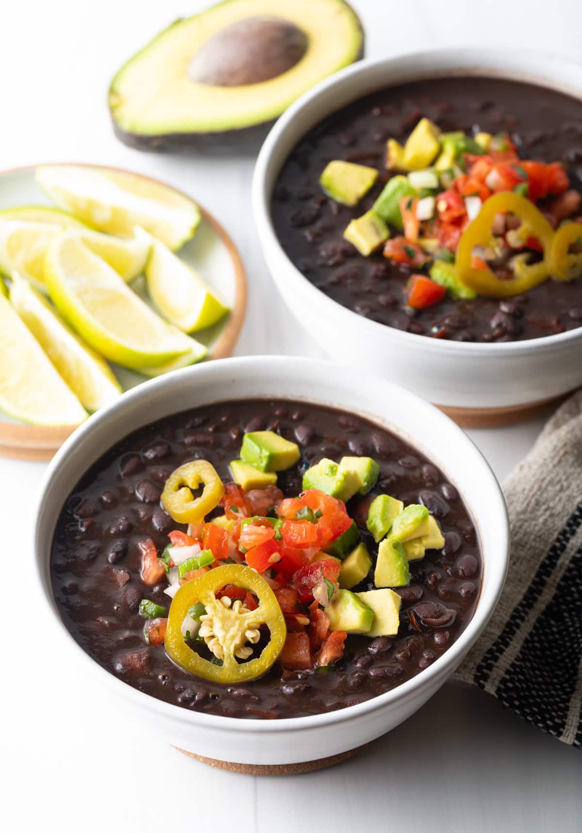 Easy Black Bean Soup Recipe - Two white bowls of black bean soup topped with slices of jalapenos, chopped avocado, and pico de gallo.