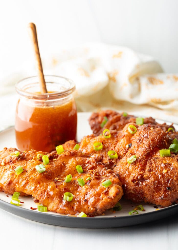 Three pieces of fried hot honey chicken on a plate. Chicken is sprinkled with sliced green onions, a jar of honey with a wood spoon in it is in the background.