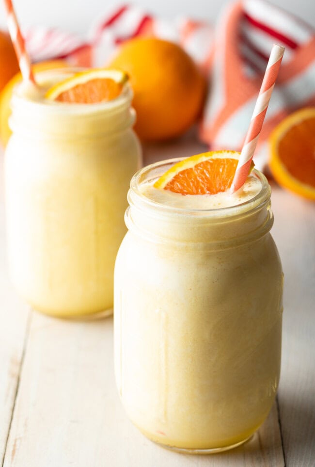Two jars filled with orange julius smoothie with straws and a fresh orange on top.