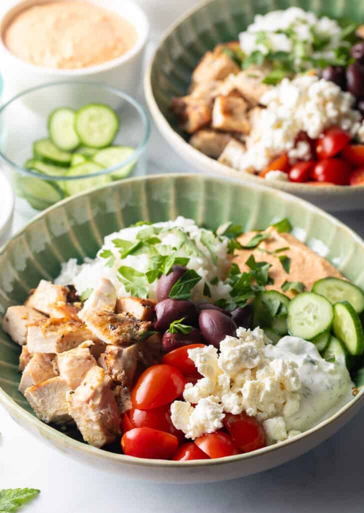 Greek chicken bowl with pieces of chicken, rice, sliced cherry tomatoes, kalamata olives, tzatziki sauce, cucumbers and feta cheese.