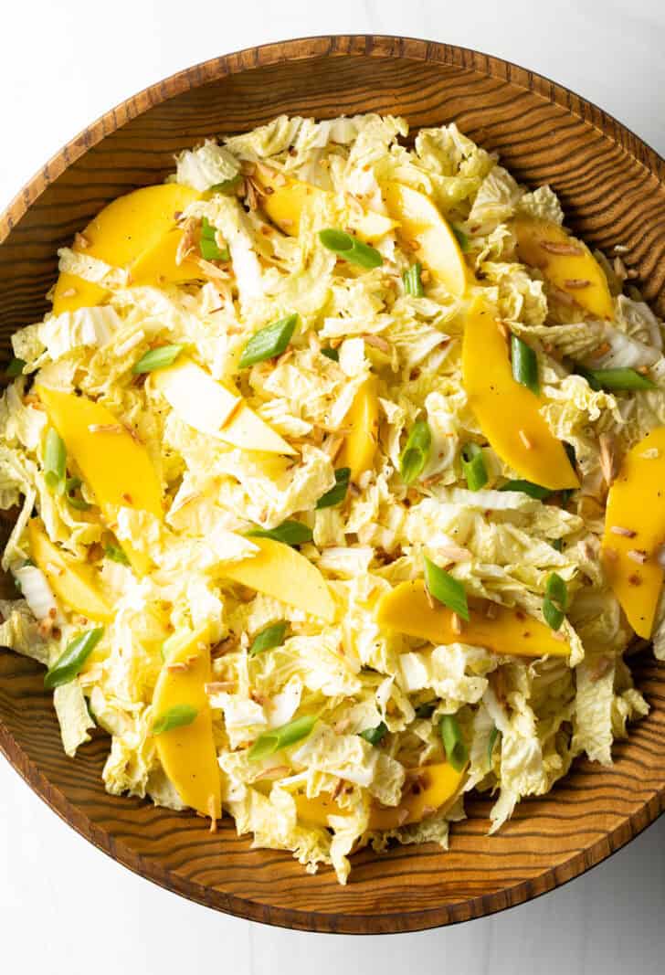 Top down view wood bowl with green napa cabbage, mango slices, and green onions.