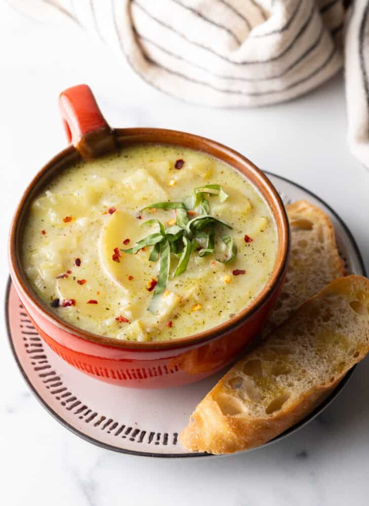 Red oversized mug filled with leek and potato soup, on a plate with slice of baguette on the side.