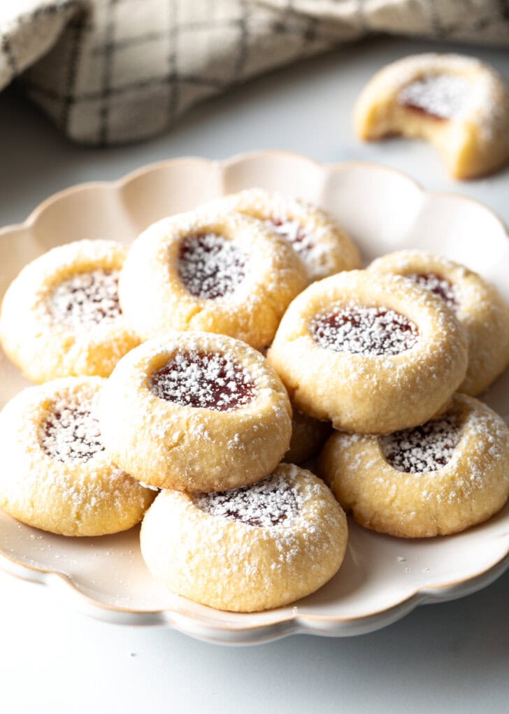 Several easy raspberry thumbprint cookies stacked two-high on a white plate, dusted with powdered sugar.