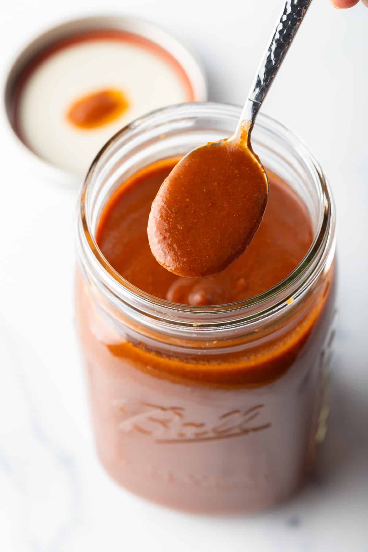 Spoonful of red enchilada sauce being held to camera over a large jar.