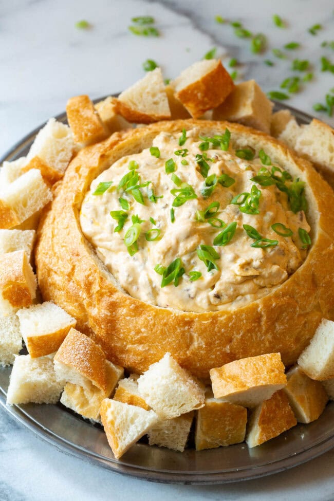 A bread bowl filled with sausage cheese dip on a plate with bread pieces for dipping.