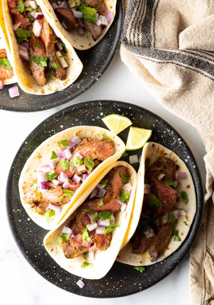 Top down view of a black plate with 3 tacos, featuring marinated steak and toppings.