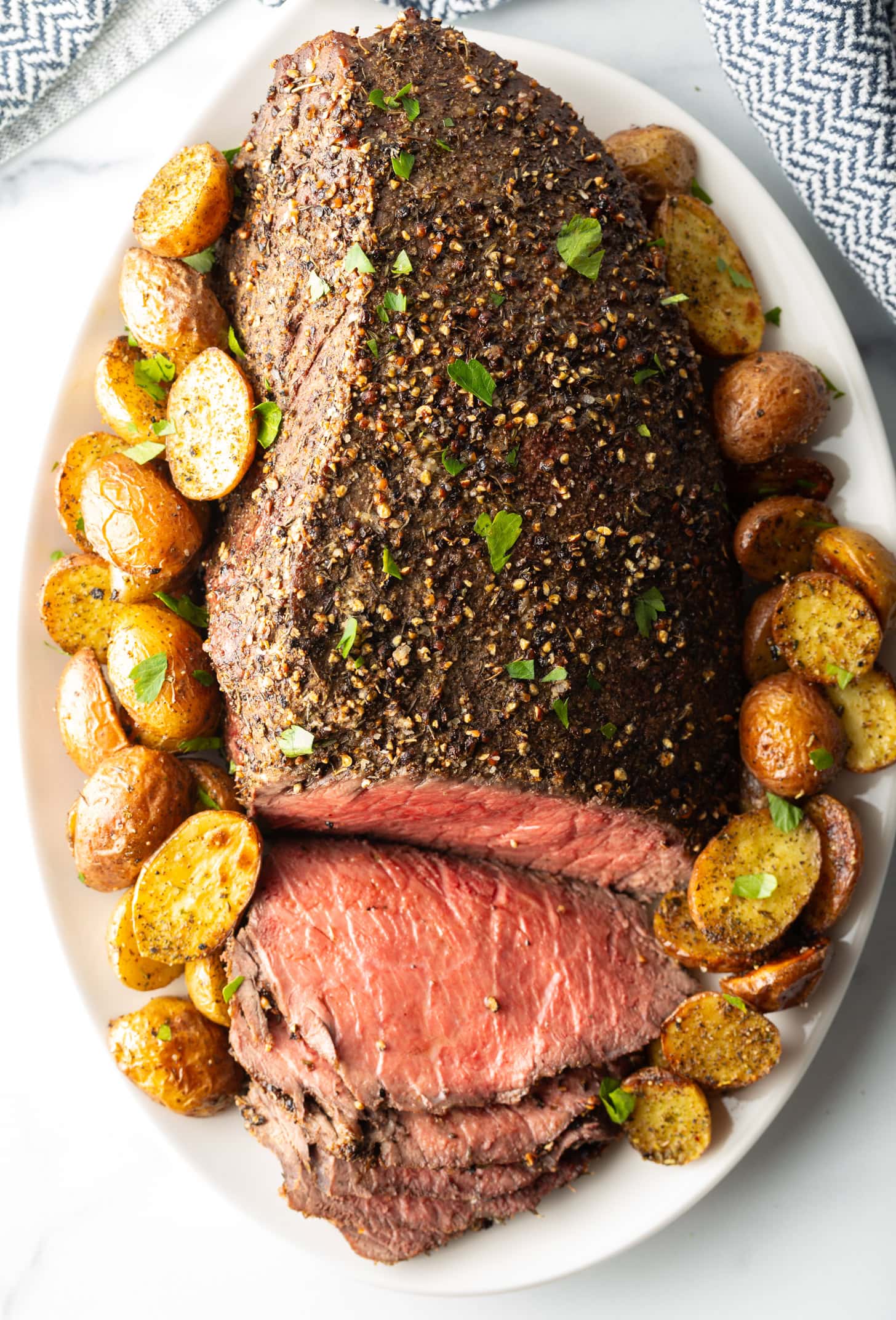 Cooked beef round roast on a metal baking pan. The front half of the roast has been thinly sliced and the meat is surrounded by roasted baby potatoes.