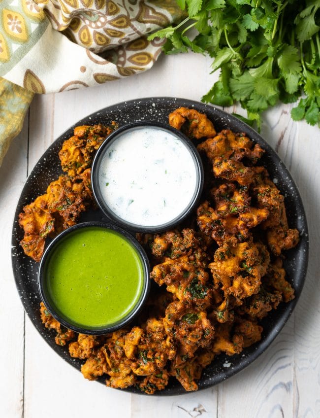 Top down view plate with kale vegetable pakora, with a ramekin of white sauce and ramekin of green sauce.